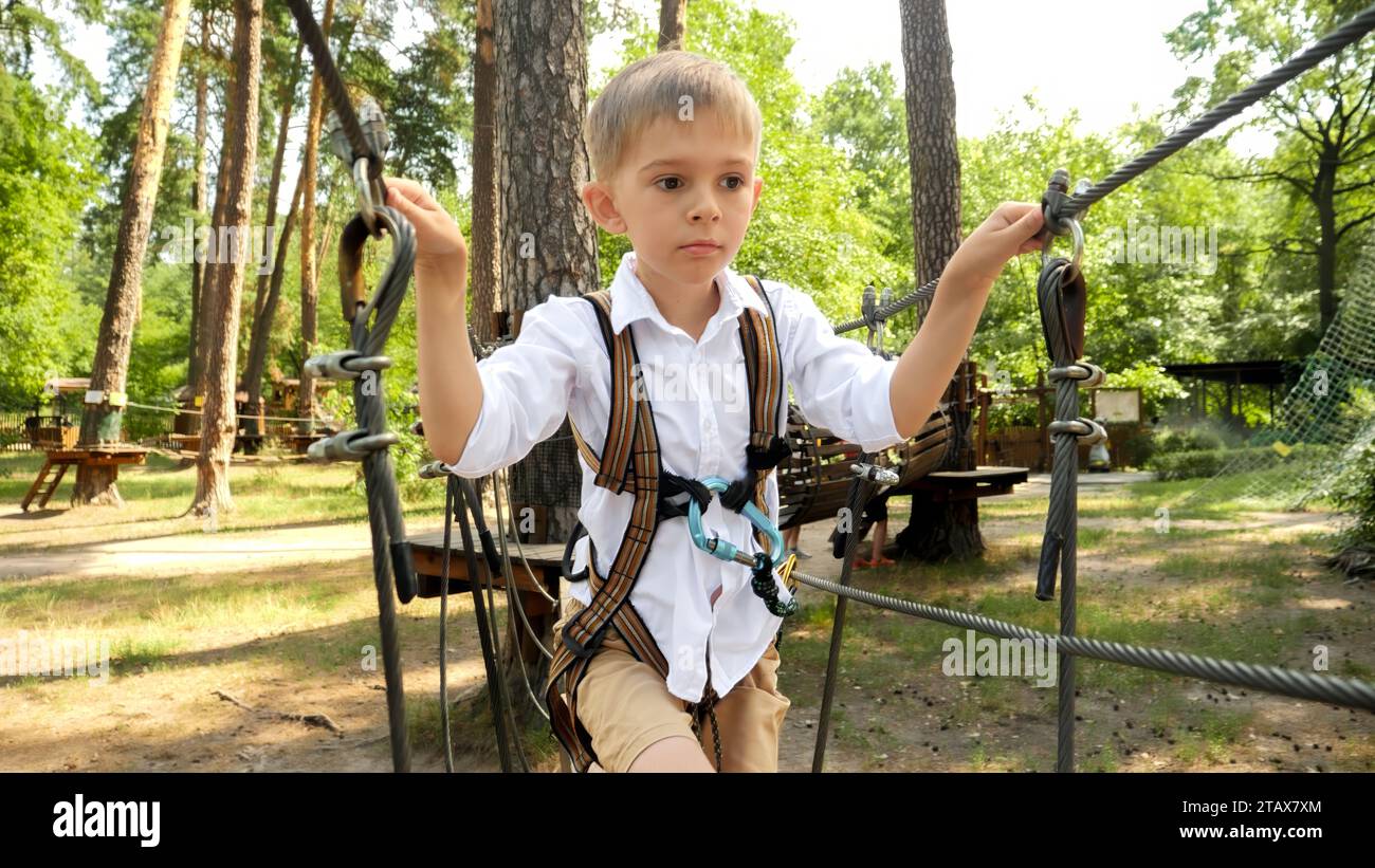 Portrait of little boy crossing the rope bridge between two trees in park. Stock Photo