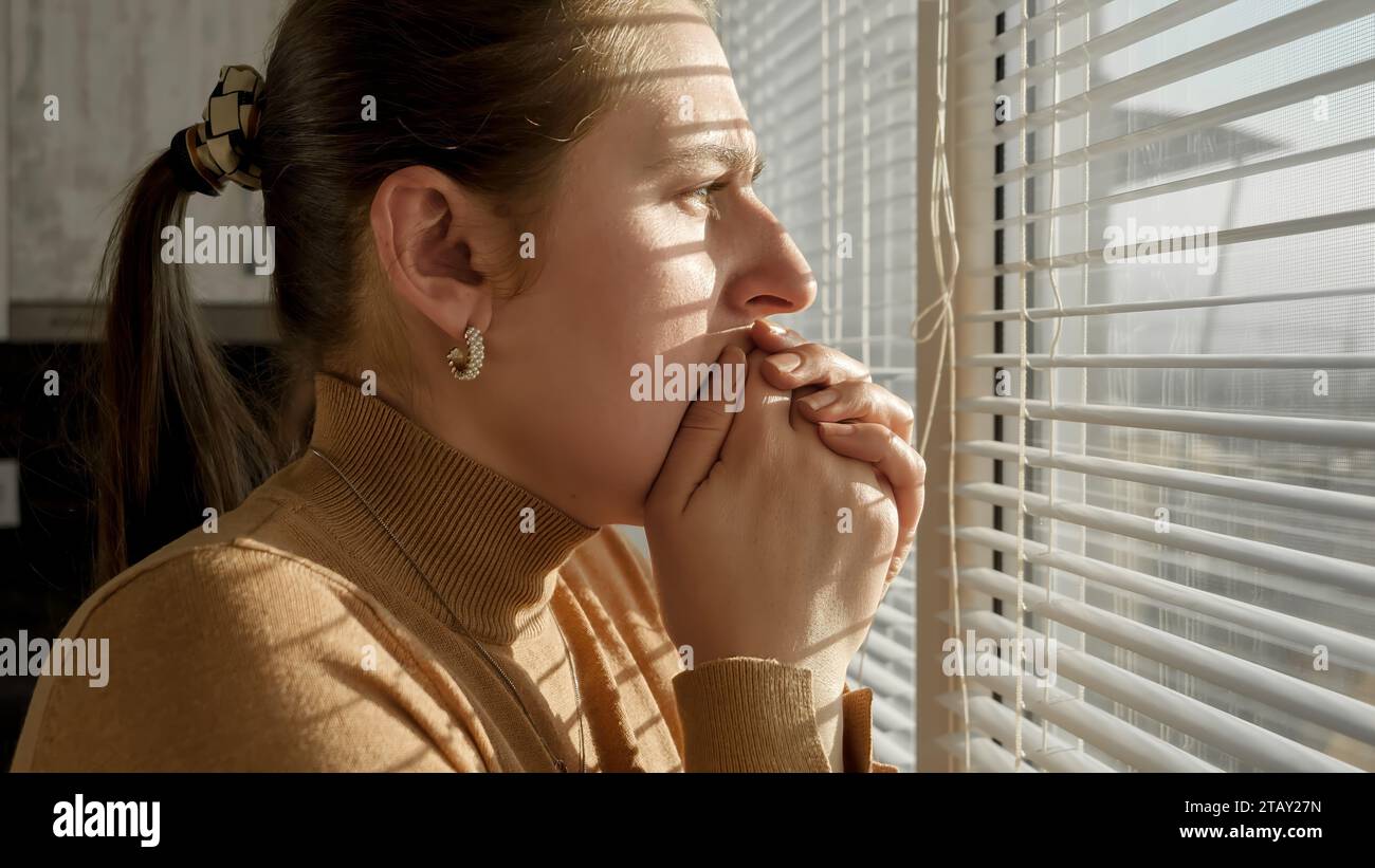 Afraid and scared woman closing her mouth with hand and looking out of the window through blinds. Stock Photo