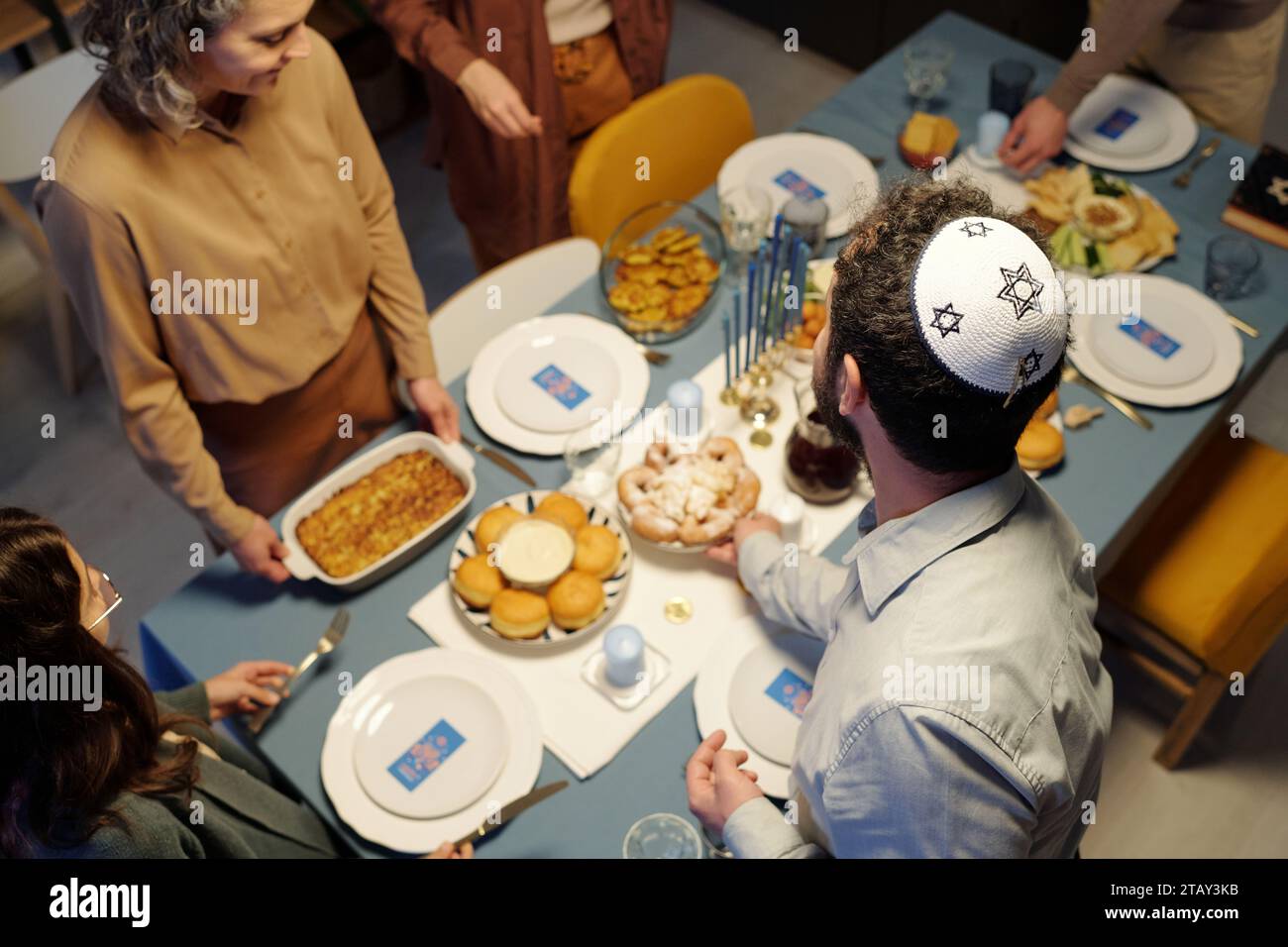 High angle of Jewish man wearing white kippah with embroidered star of David putting plate with homemade snack on served table Stock Photo