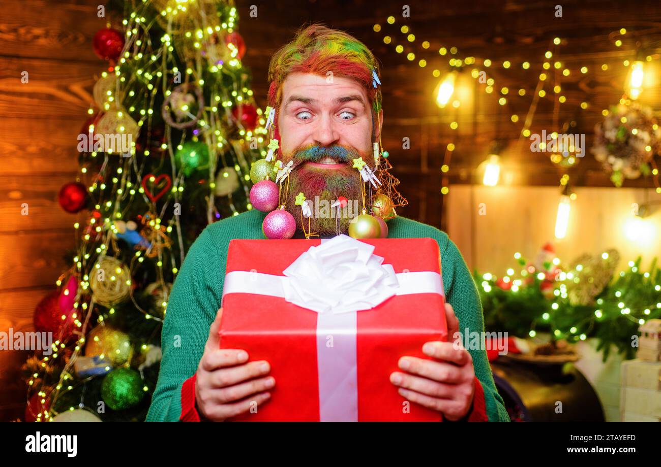 Funny bearded man with Christmas gift box. Smiling man with decoration balls in his beard with New year present. Bearded man with Christmas decorated Stock Photo