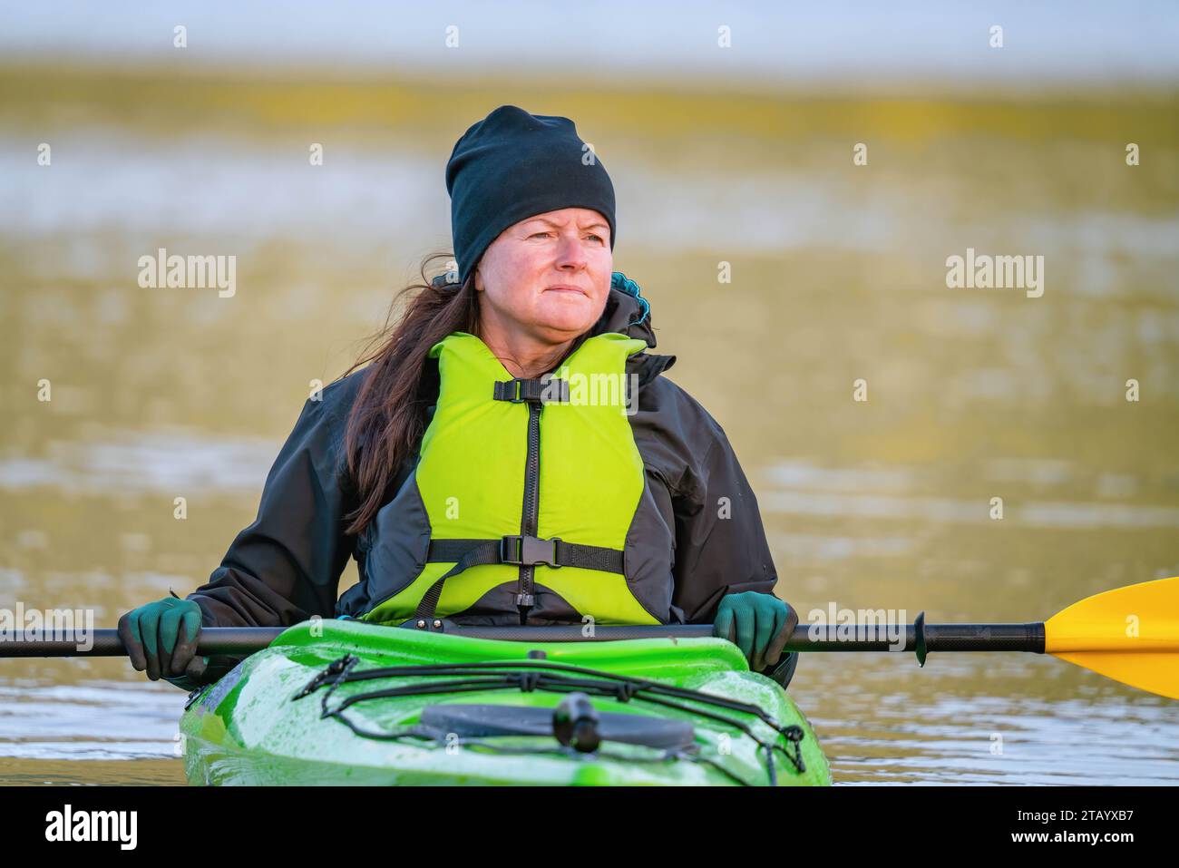 Mature women in green safety life jacket sits in green kayak and looks aside holding yellow paddle across boat. Very close up photo on still water wit Stock Photo