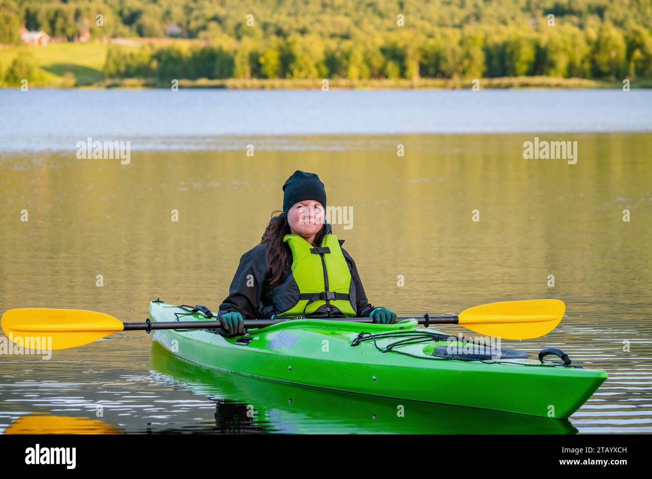 Mature women in green safety life jacket sits in green kayak and look at camera holding yellow paddle across boat. Very close up photo on still water Stock Photo