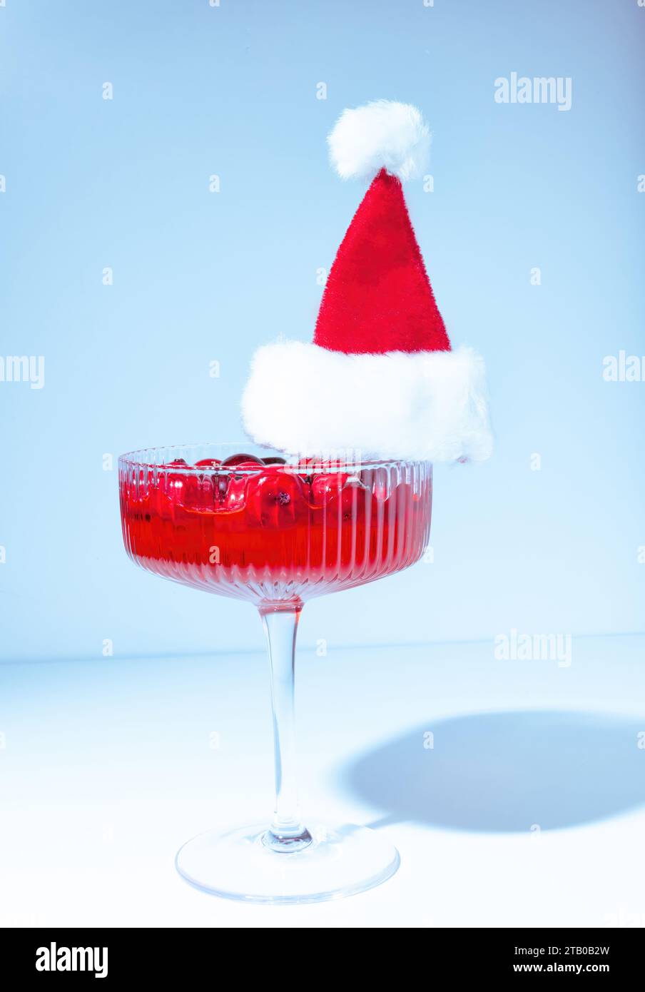 A Holiday Red Cranberry Cocktail in a Coupe Champagne Glass W earring a Furry Red Santa Claus Hat on a Blue Pastel Background Stock Photo