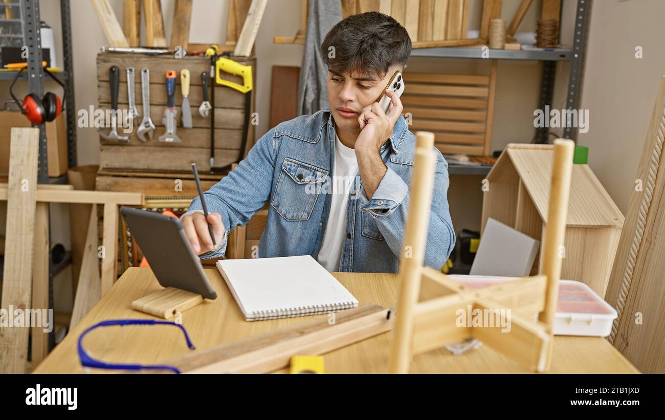 Experienced young hispanic carpenter multitasking, talking on a smartphone while using a touchpad in a busy carpentry workshop Stock Photo