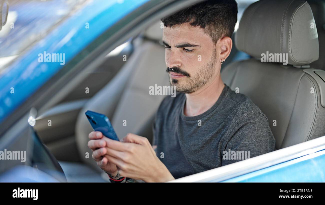 Young hispanic man using smartphone sitting on car at street Stock Photo