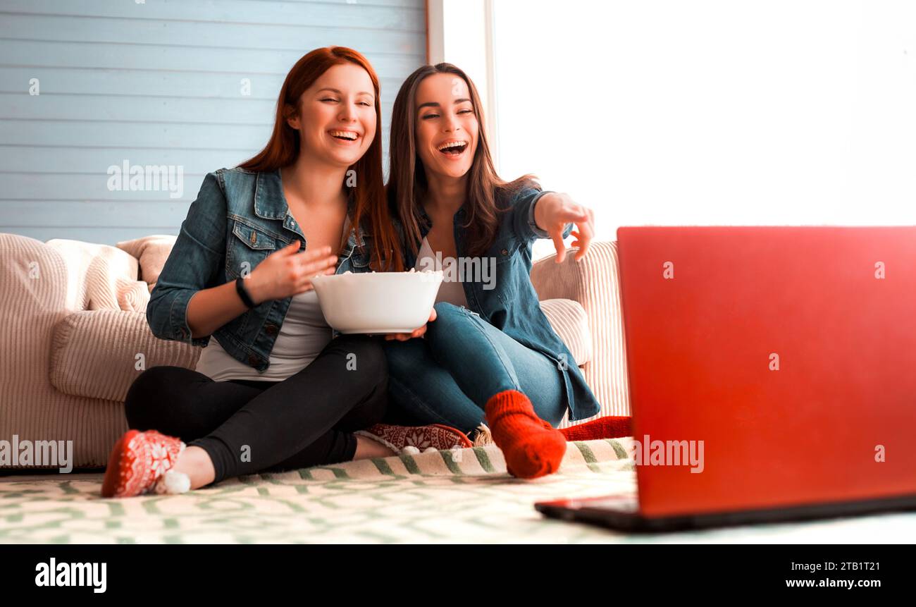 Close up photo of girl friends sitting on the floor near the couch in front of laptop and they watch a movie and eat popcorn. Funny friends relaxing t Stock Photo