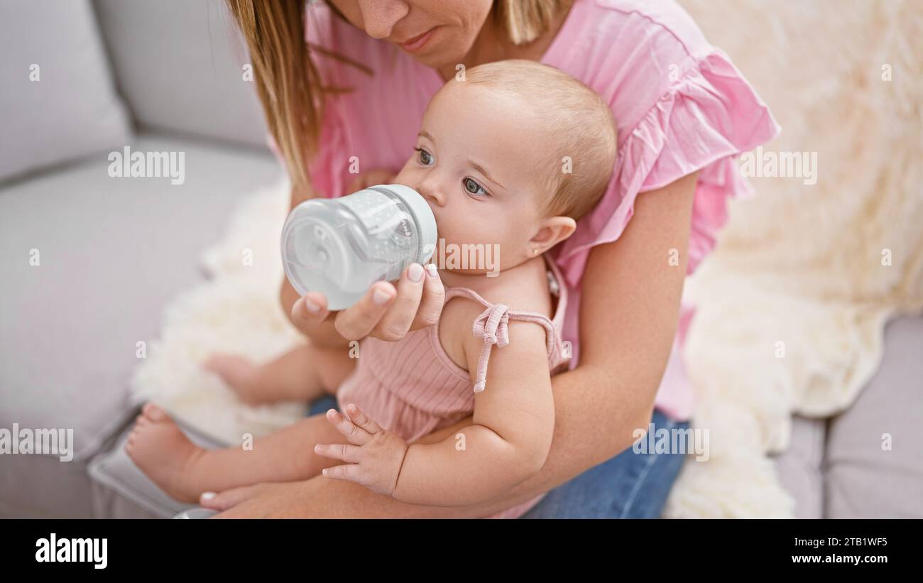Loving mother giving a milk feeding bottle to her baby daughter while sitting in the relaxed comfort of their family home, cherishing an intimate indo Stock Photo