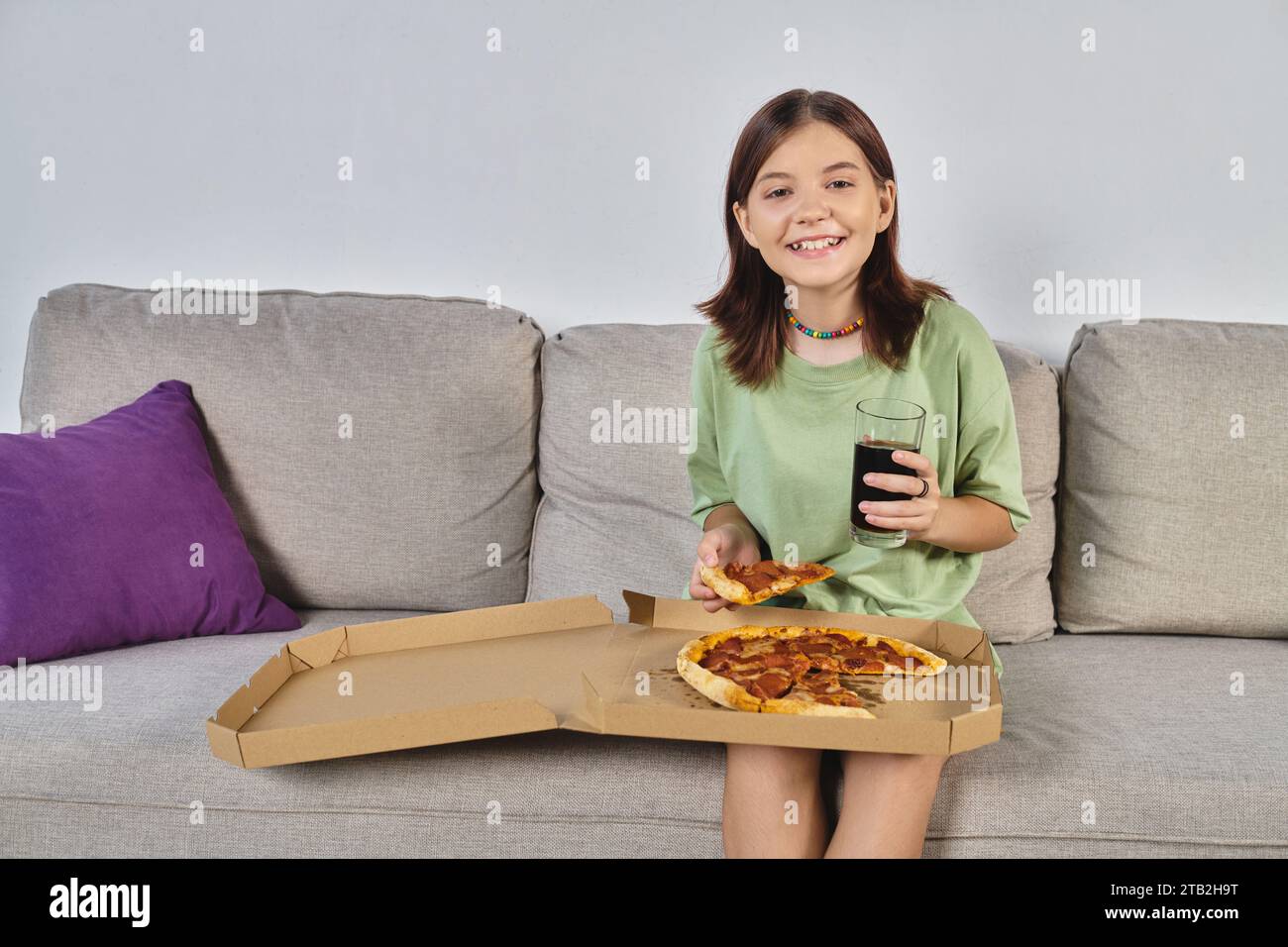 joyful teenage girl looking at camera while sitting on couch with pizza and soda, meal time Stock Photo