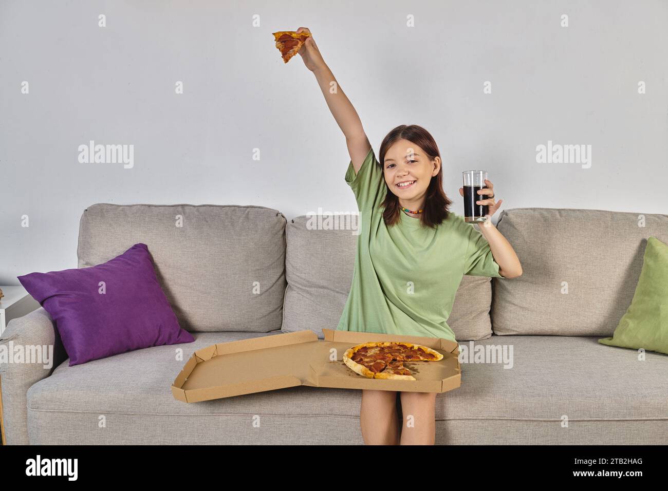 excited teenage girl sitting on couch with pizza and glass of cola and looking at camera, meal time Stock Photo