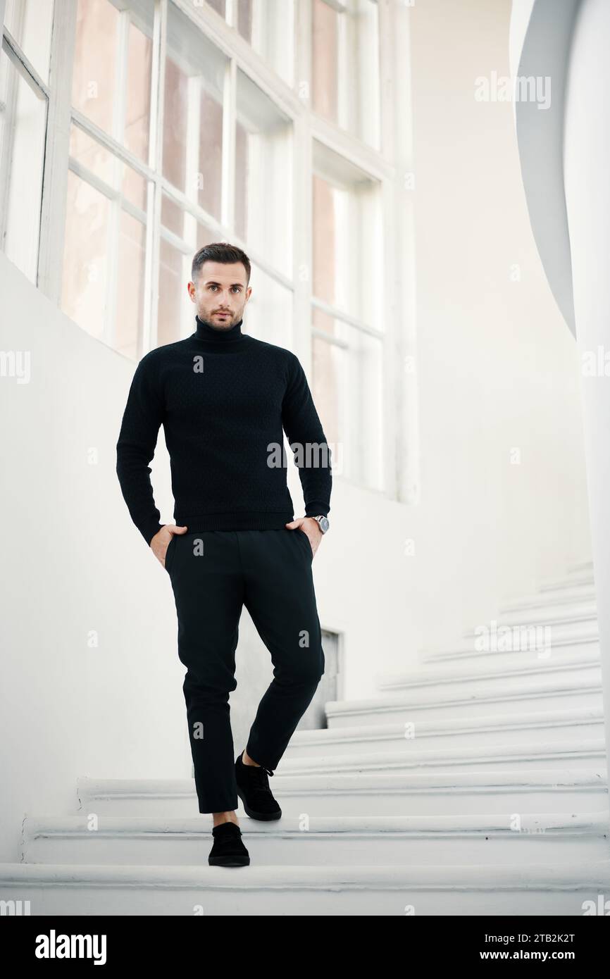 Fashionable man in a sleek black turtleneck descends a white modern staircase, exuding confidence and style. Stock Photo