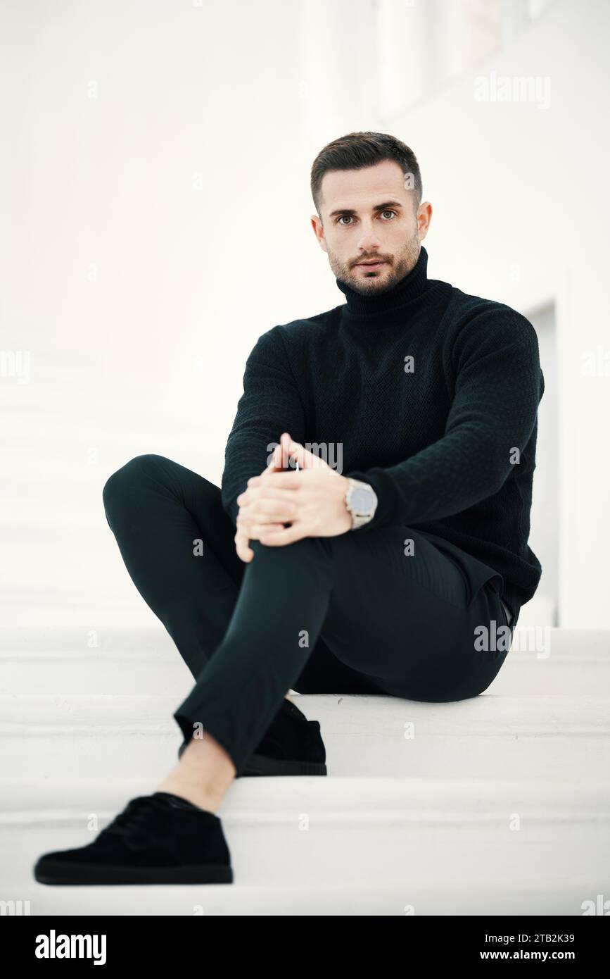 Fashionable man model sits with confidence on white staircase, dressed in all black clothing Stock Photo