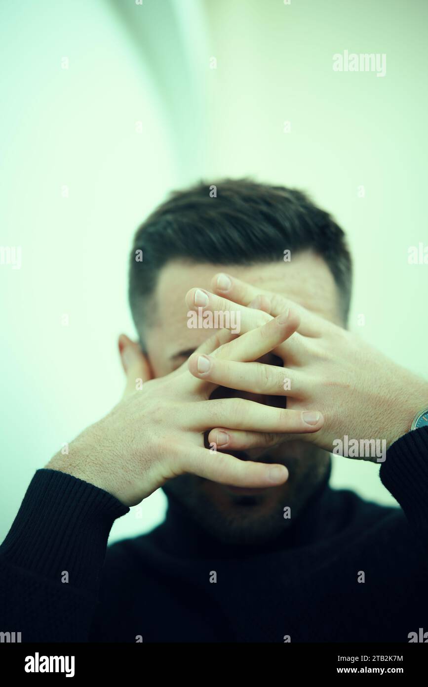 Young man in a dark turtleneck covers his face with his hands in a thoughtful or emotional moment. People emotions concept Stock Photo