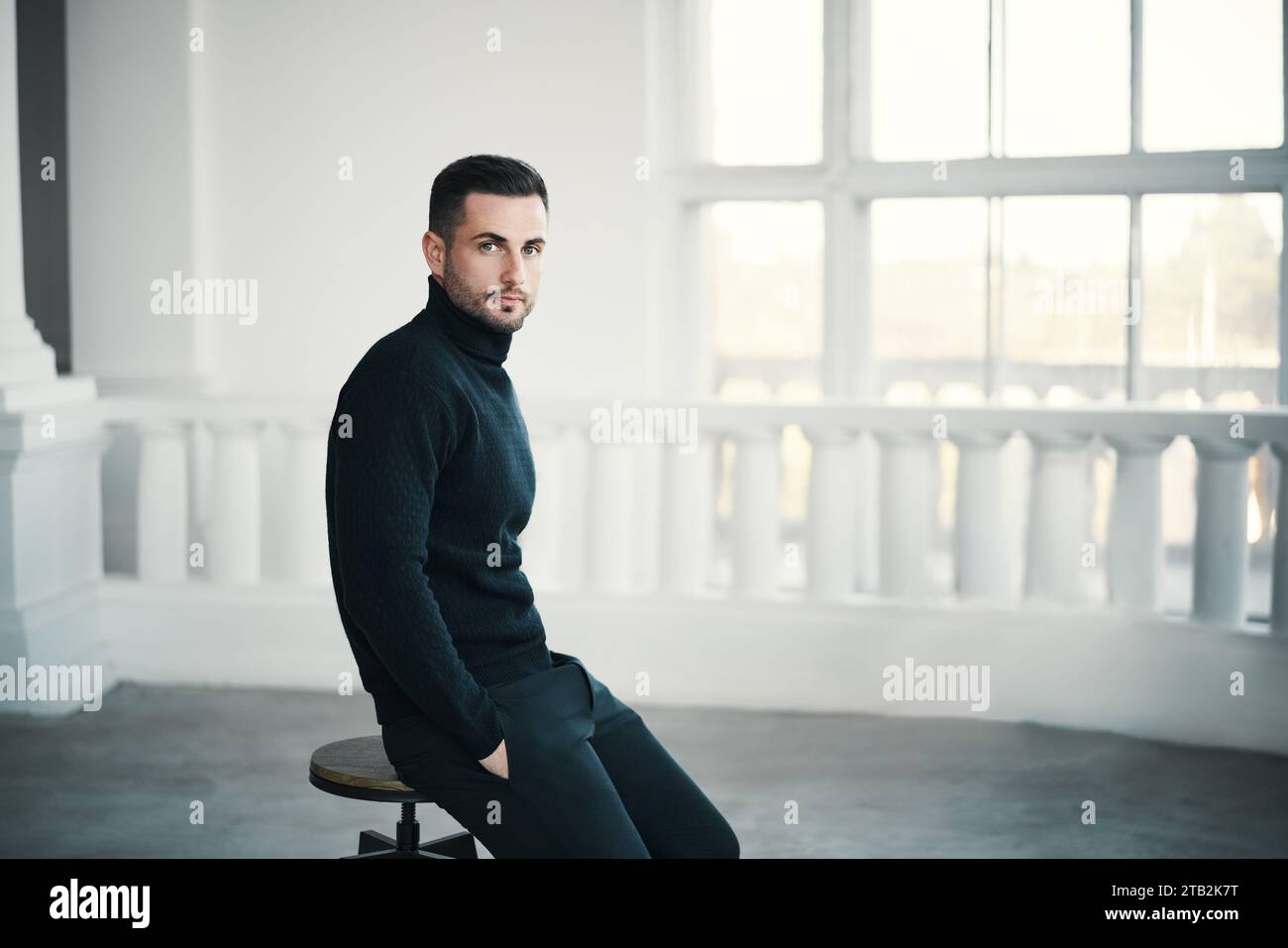 Trendy handsome man sits thoughtfully on chair in spacious white room. Male beauty Stock Photo