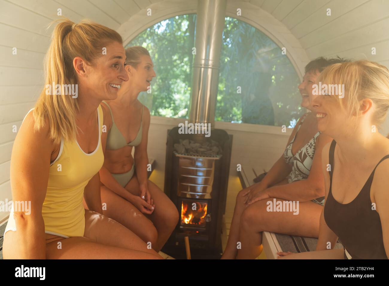 Four women enjoying a conversation in a brightly lit Finnish barrel sauna with a stove. Stock Photo