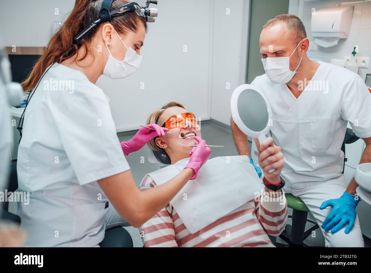 Smiling female lying in stomatology chair, gazing at mirror, female doctor  with excavator and mirror medical tools and assistant man after teeth whit Stock Photo