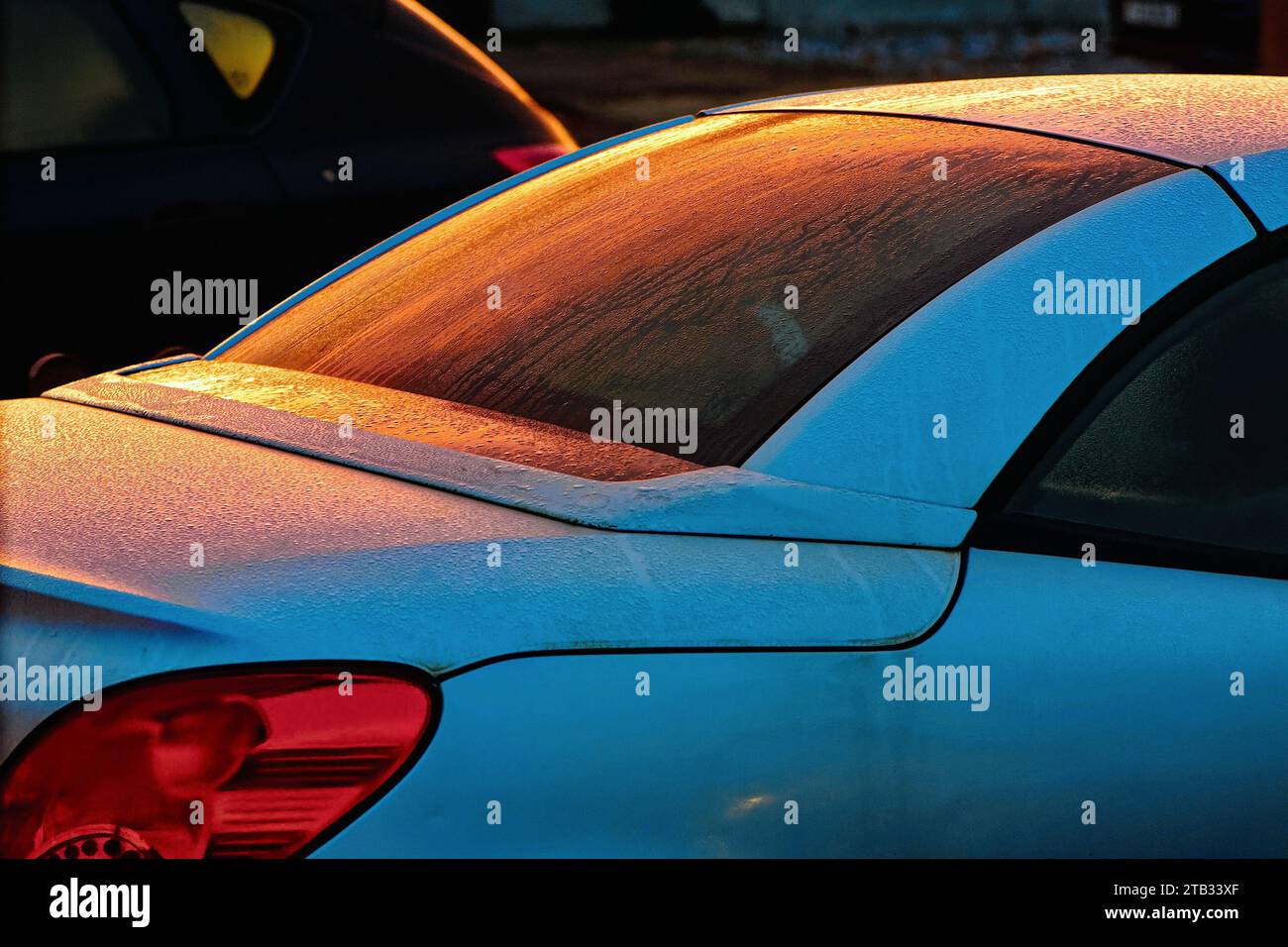 Close up of a silver car with reflections of red dawn light on rear window covered in frost and raindrops creating an abstract shape Stock Photo