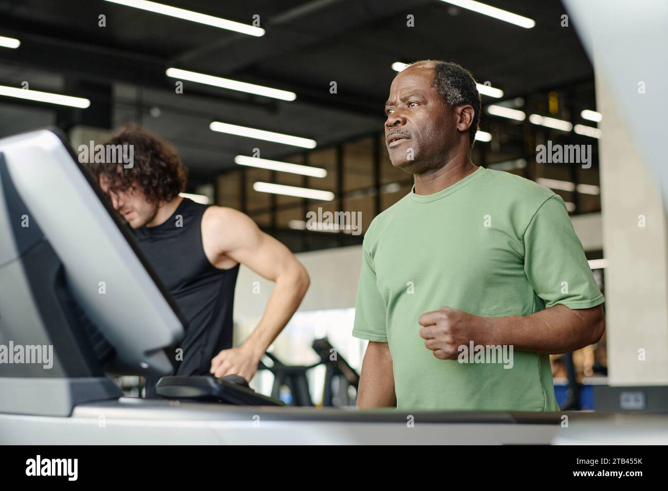 Elderly black man standing on treadmill at gym with caucasian gym goer ...