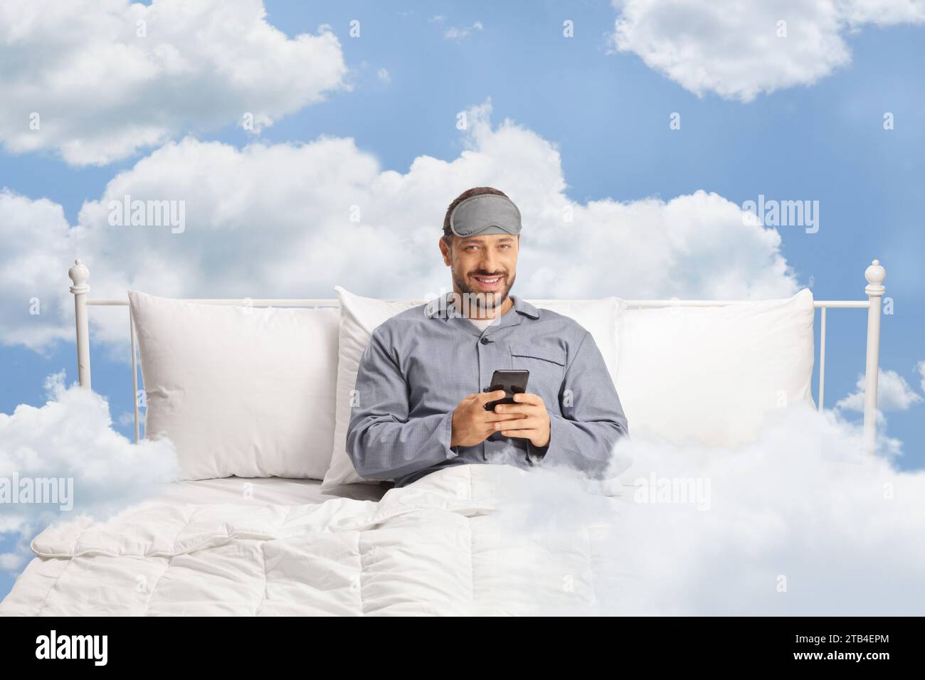 Man in pajamas sitting in a bed up in the sky and holding a smartphone Stock Photo