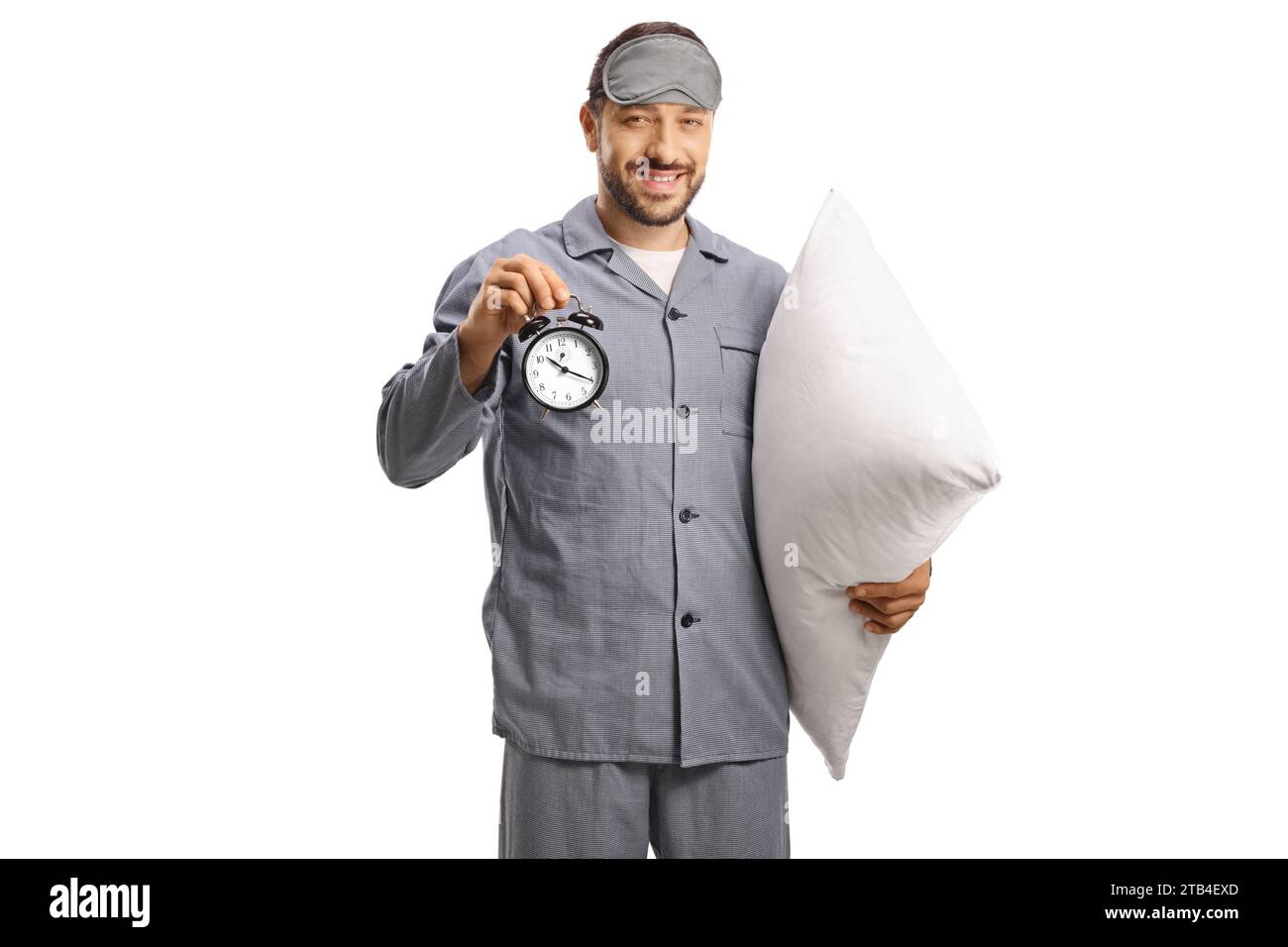 Man in pajamas with a sleeping mask holding a pillow and an alarm clock isolated on white background Stock Photo