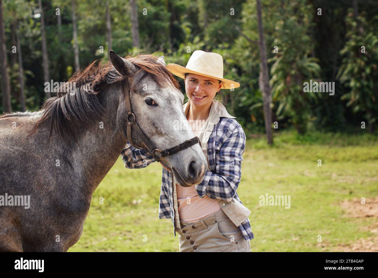 Young woman in shirt and straw hat posing next to gray horse, jungle trees background - horseriding, ranch at Isalo Park, Madagascar Stock Photo