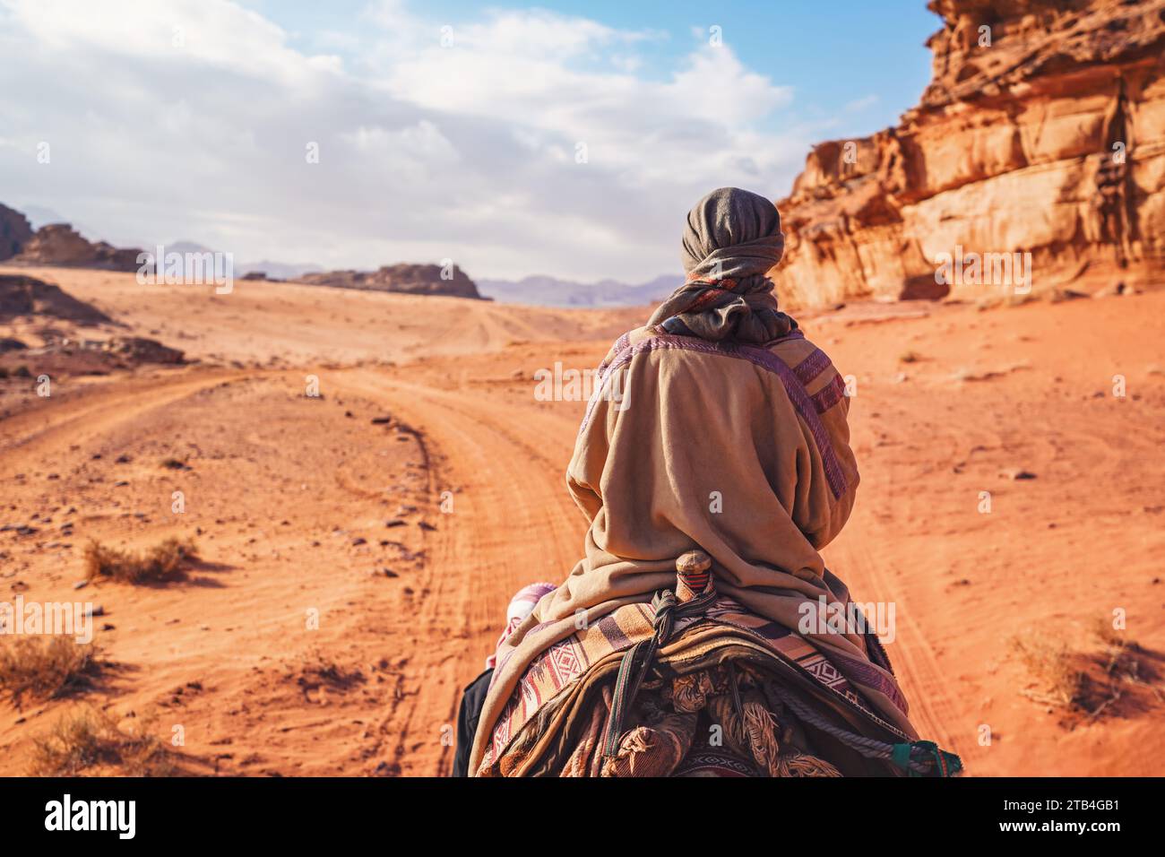 Young woman riding camel in desert. It's quite cold so she is wearing traditional Bedouin coat - bisht - and head scarf, rocky hills at side Stock Photo