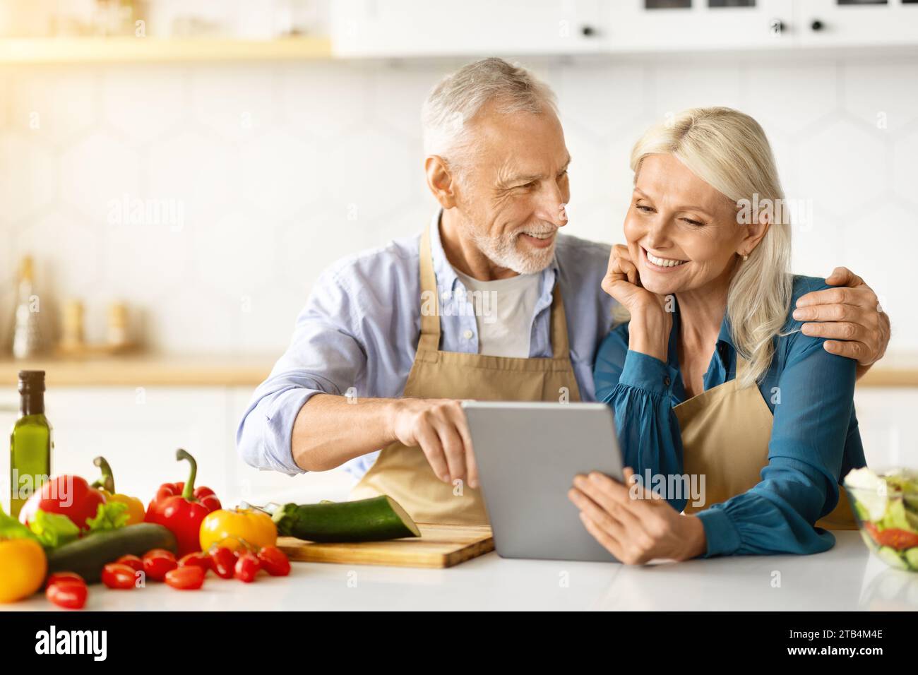 Cheerful Elderly Couple Using Digital Tablet While Cooking Healthy Lunch In Kitchen Stock Photo