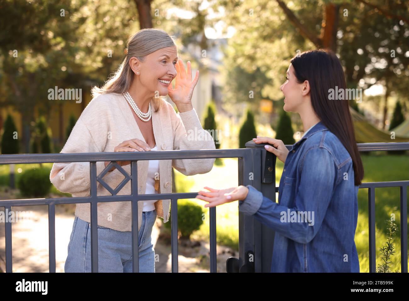 Friendly relationship with neighbours. Happy women talking near fence outdoors Stock Photo