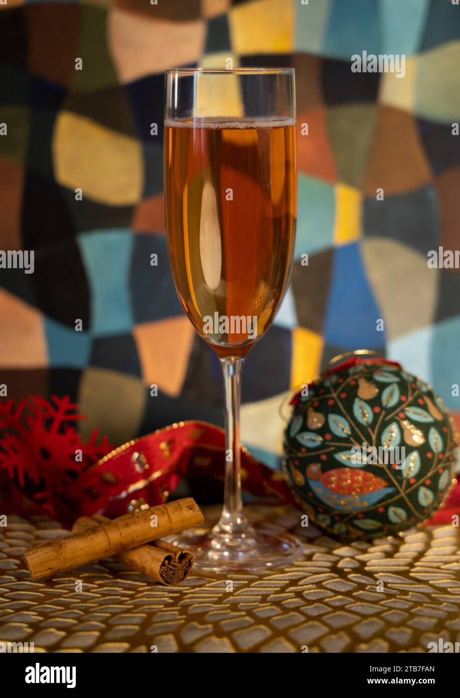 Tall glass of champagne with Christmas decorations including two cinnamon sticks, a green bauble, a red snowflake in front of a painting Stock Photo