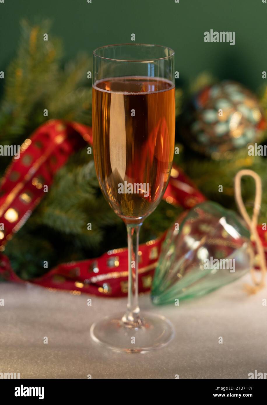 Tall glass of champagne in front of green background with Christmas tree foliage, glass baubles with a red ribbon on white fabric Stock Photo
