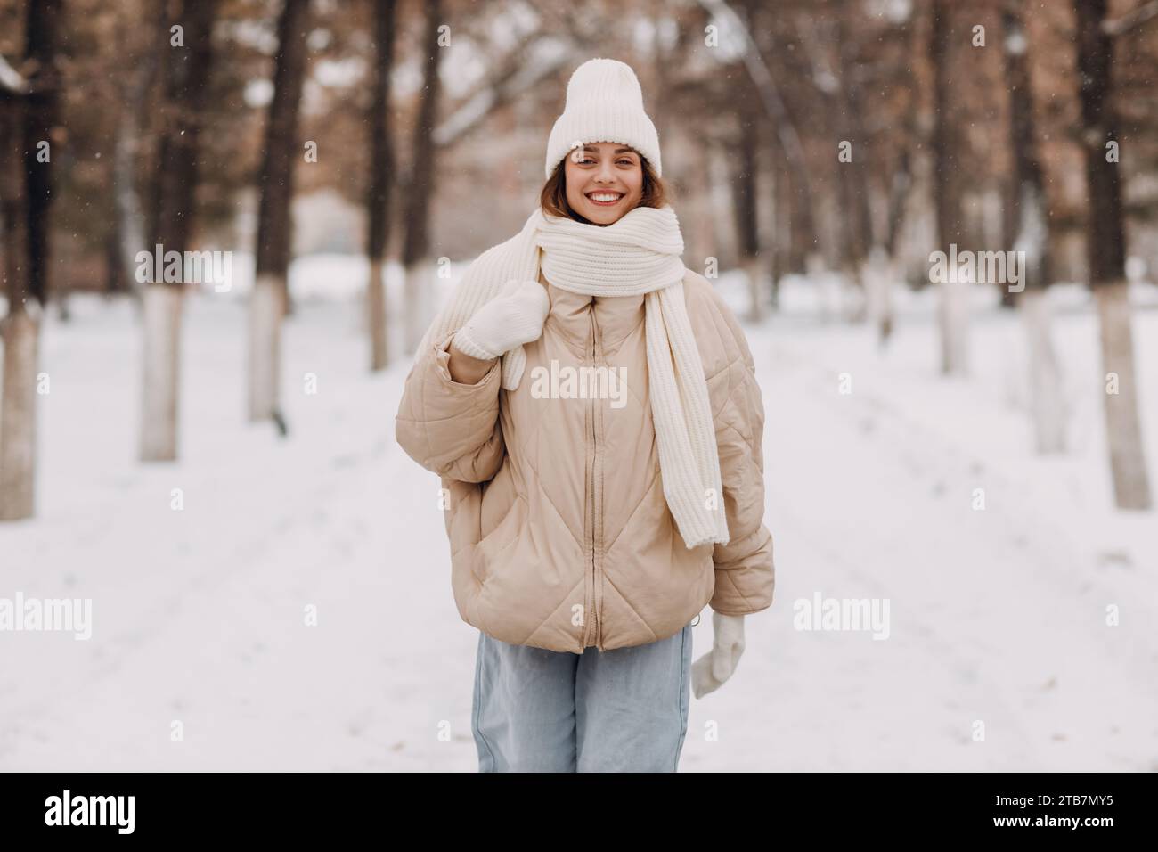 Happy smiling young woman dressed down jacket scarf hat and mittens enjoys winter weather and walking through the snowy winter park Stock Photo