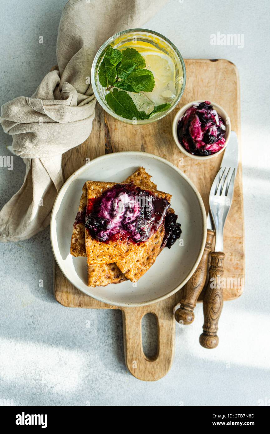 Freshly made crepes are served with a dollop of sour cream and a generous spread of blueberry jam on a rustic wooden board. Stock Photo