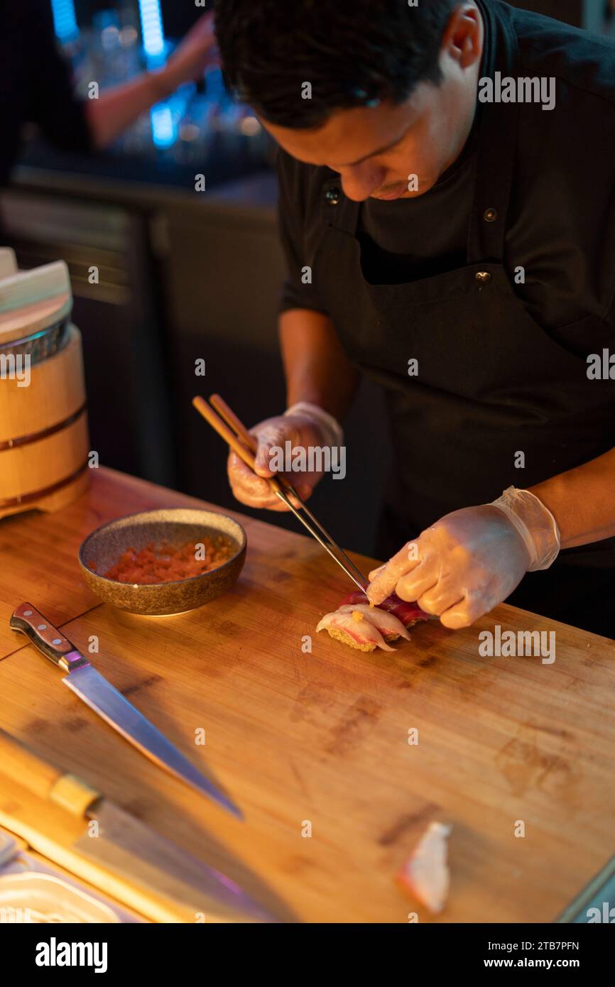 A sushi chef meticulously assembles a piece of nigiri sushi at a warmly lit restaurant counter, showcasing culinary precision. Stock Photo