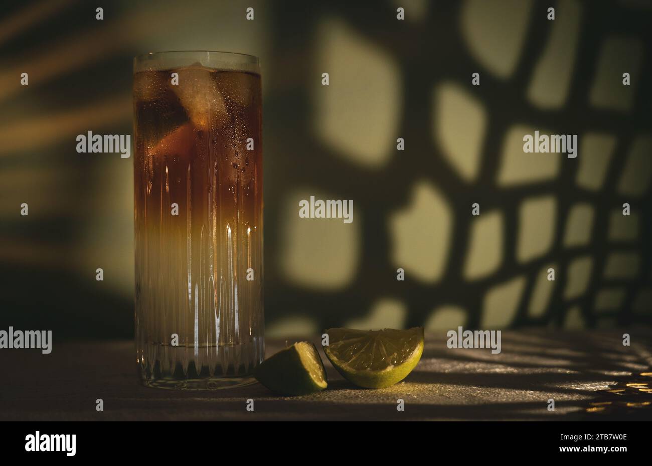 Dark and Stormy cocktail with ice and lime wedges on white table cloth in front of a green dappled background/wall Stock Photo
