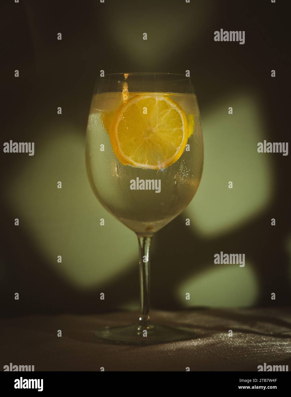 Gin and tonic cocktail in wine glass with ice and lemon slices in front of light green wall on a white table cloth Stock Photo