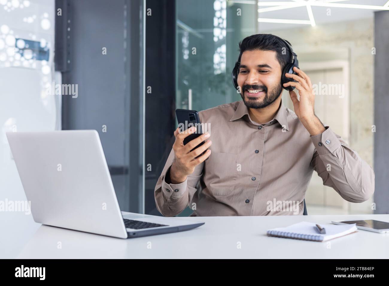 Satisfied successful hindu man at workplace inside office, man with phone in hands and headphones watching online video and listening to music audio p Stock Photo