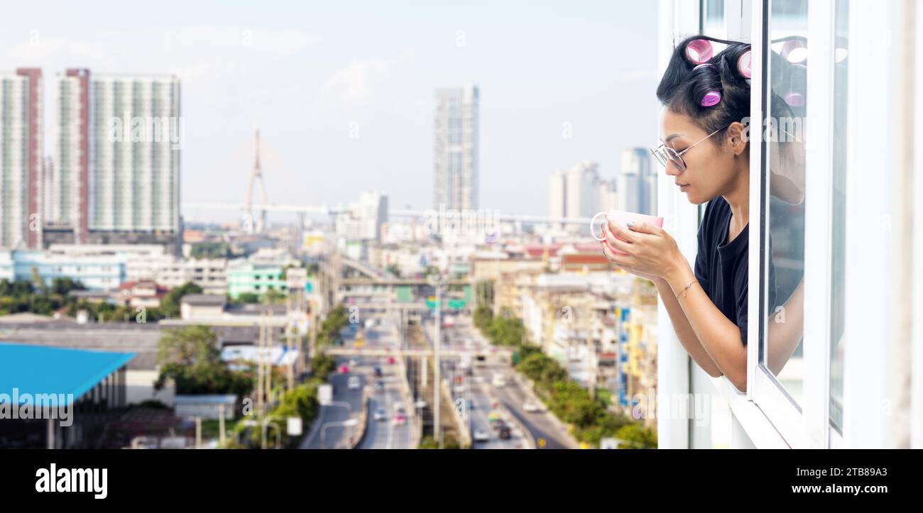 A young woman in curlers looks out of the window of a high-rise building in the city center Stock Photo