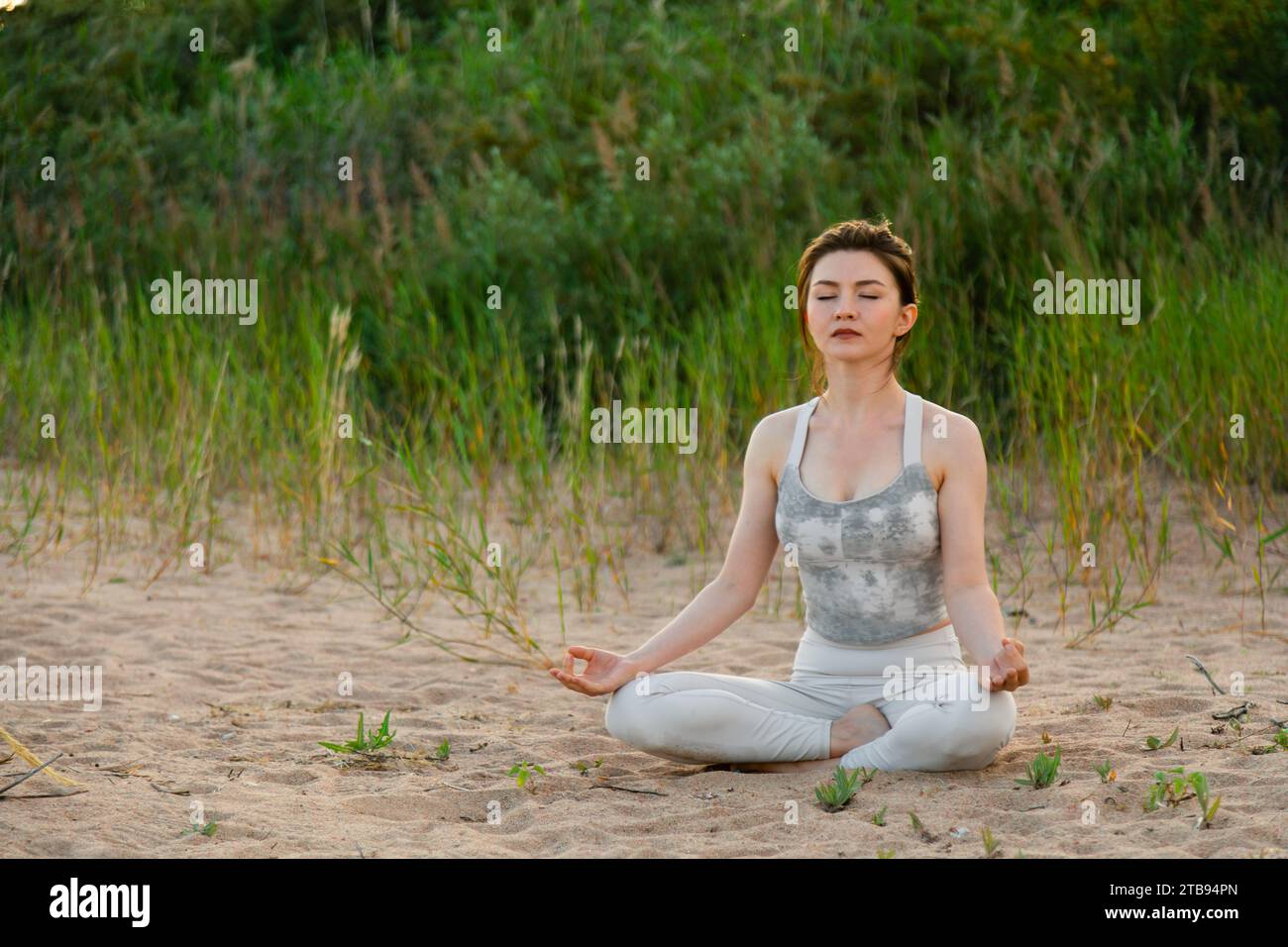 Young beautiful woman practicing yoga outdoors sitting on sand in lotus ...