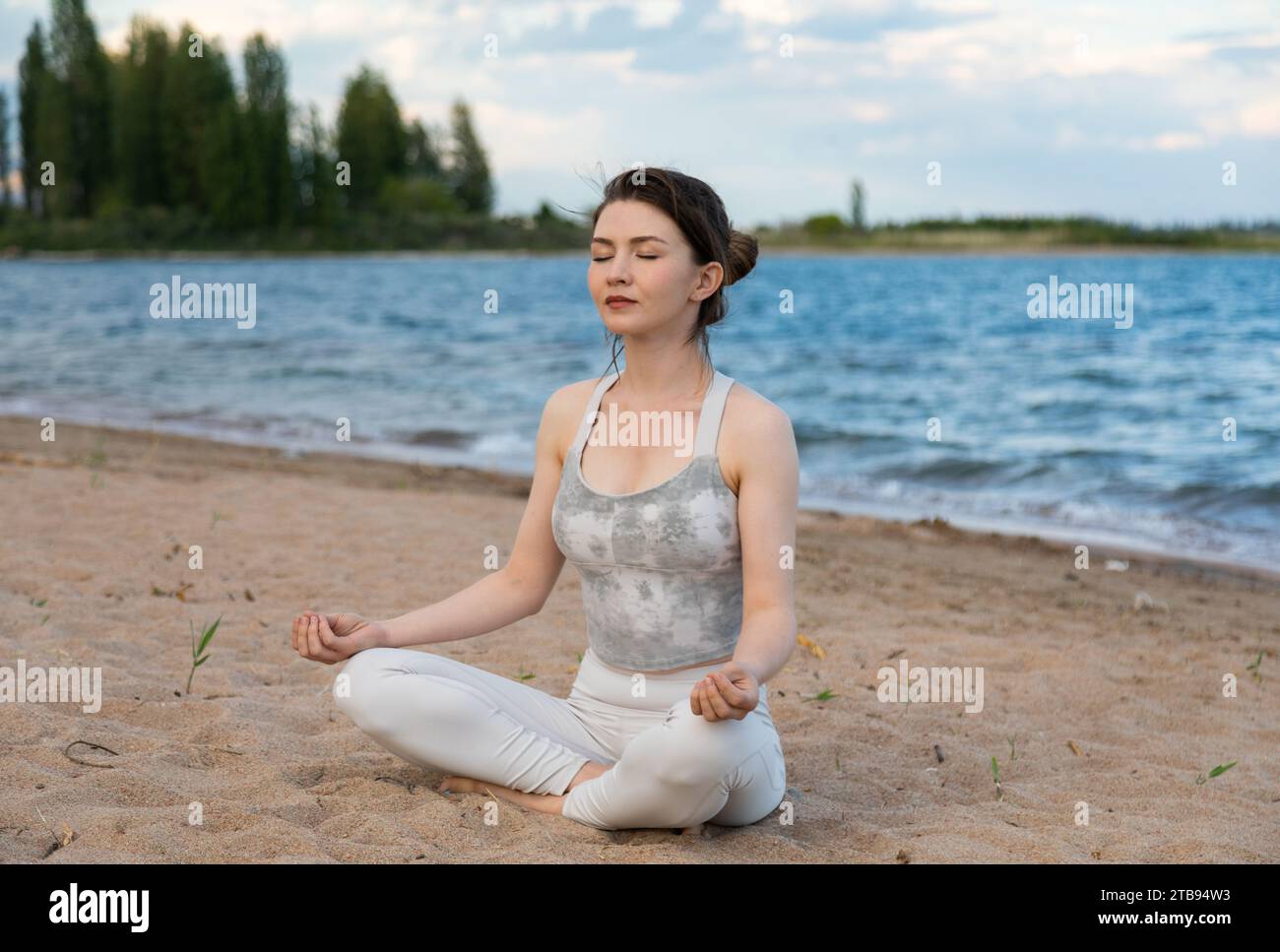 Young beautiful woman practicing yoga outdoors sitting on a lake shore ...