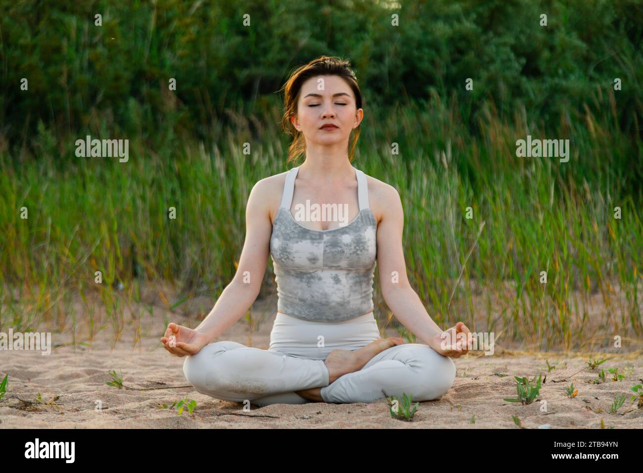 Young beautiful woman practicing yoga outdoors sitting on sand in lotus ...