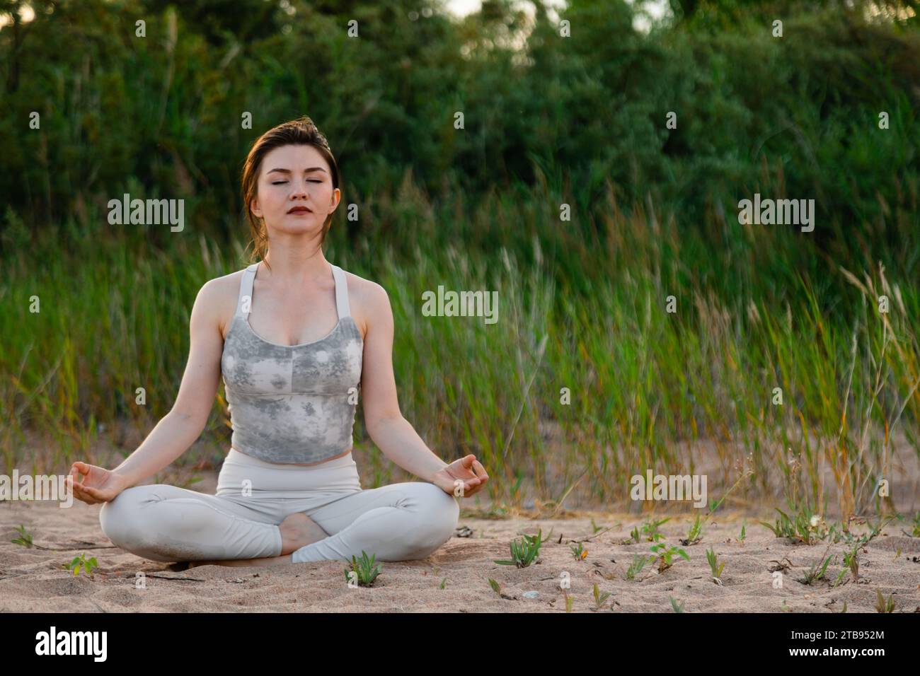 Young beautiful woman practicing yoga outdoors sitting on sand in lotus ...