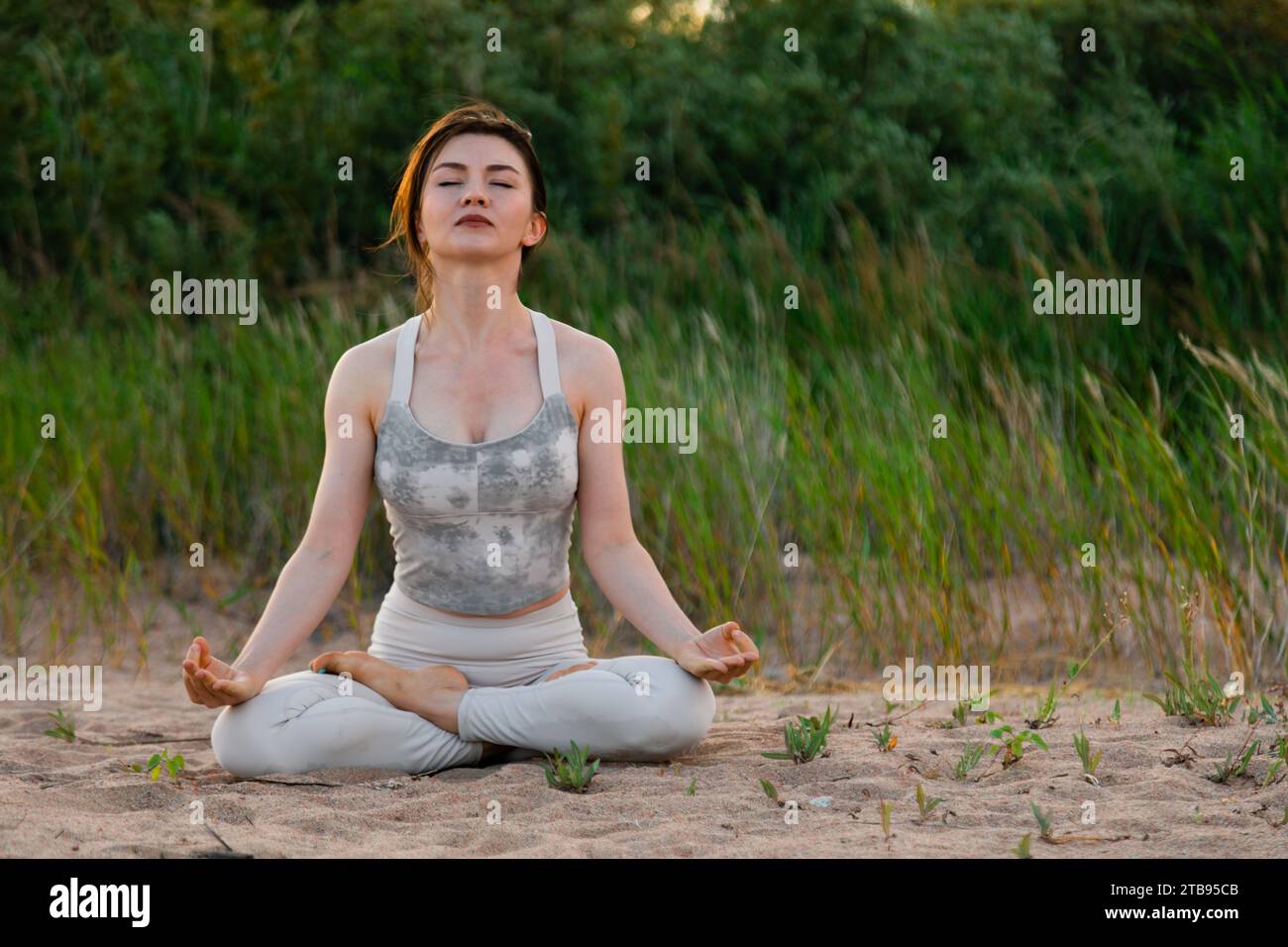 Young beautiful woman practicing yoga outdoors sitting on sand in lotus ...