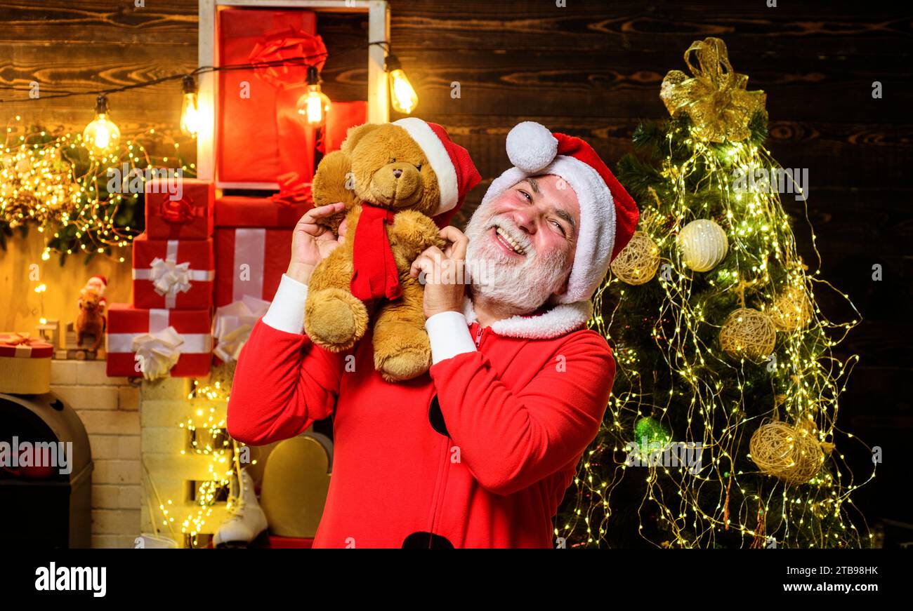 Christmas holiday. Smiling Santa Claus with toy teddy bear in room decorated for Christmas. Bearded man in Santa costume with plush teddy. Merry Stock Photo