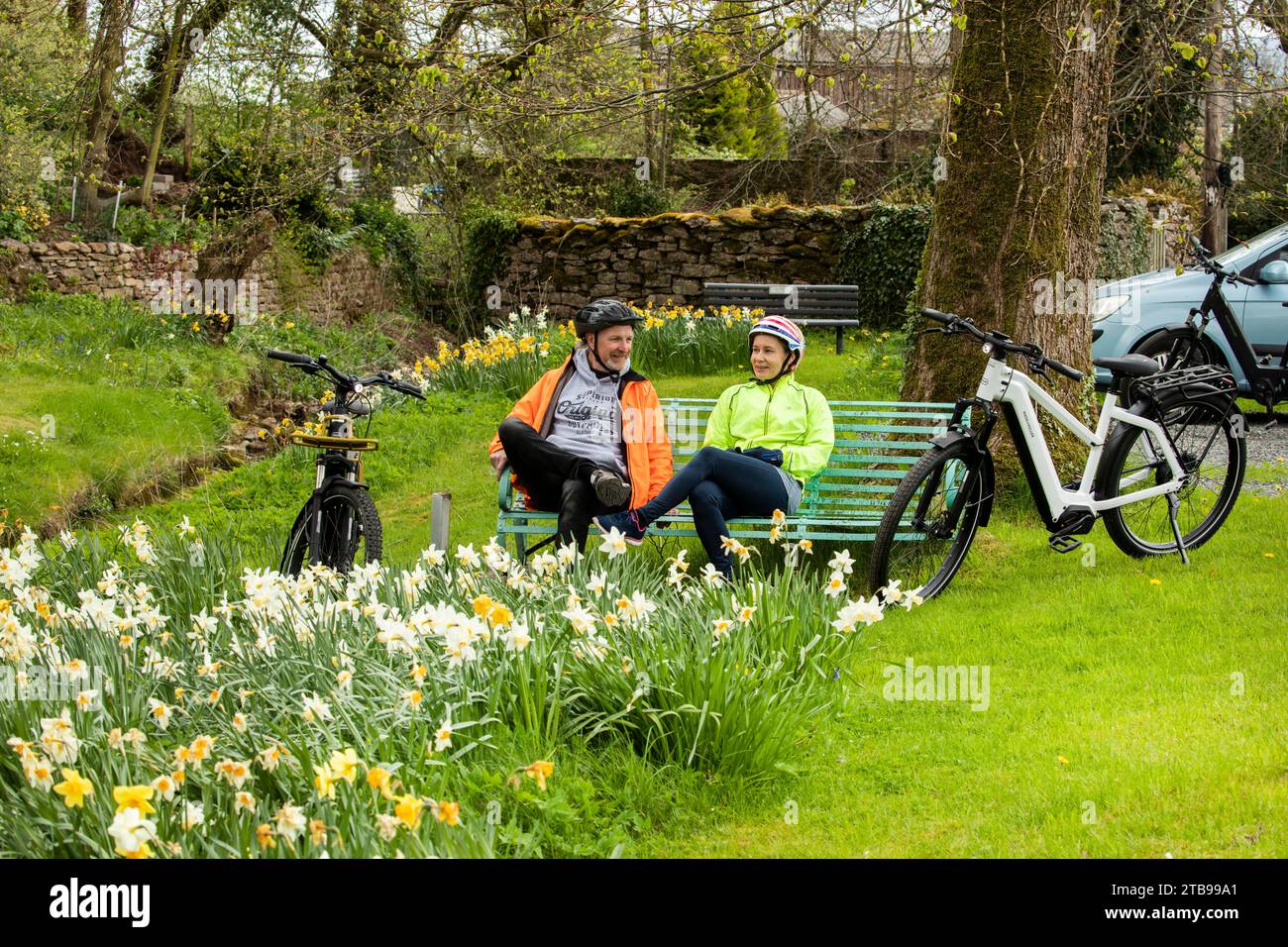 Man and woman cyclists with electric e bikes sit on bench in a pretty village in springtime, Hartley. Kirkby Stephen, North Pennines, Cumbria Stock Photo