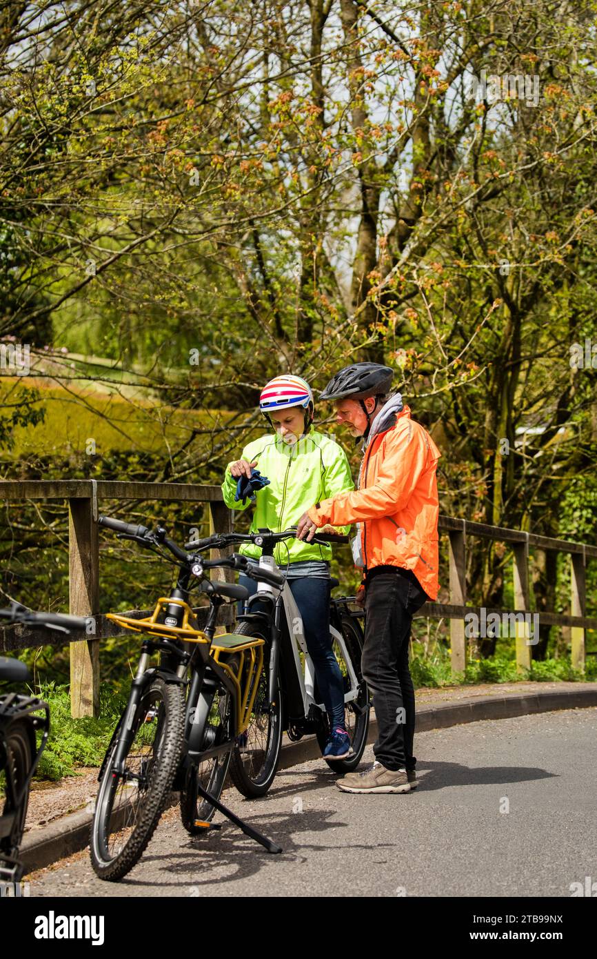 Man and woman cyclists riding electric e bikes in a pretty village in springtime, Hartley. Kirkby Stephen, North Pennines, Cumbria Stock Photo