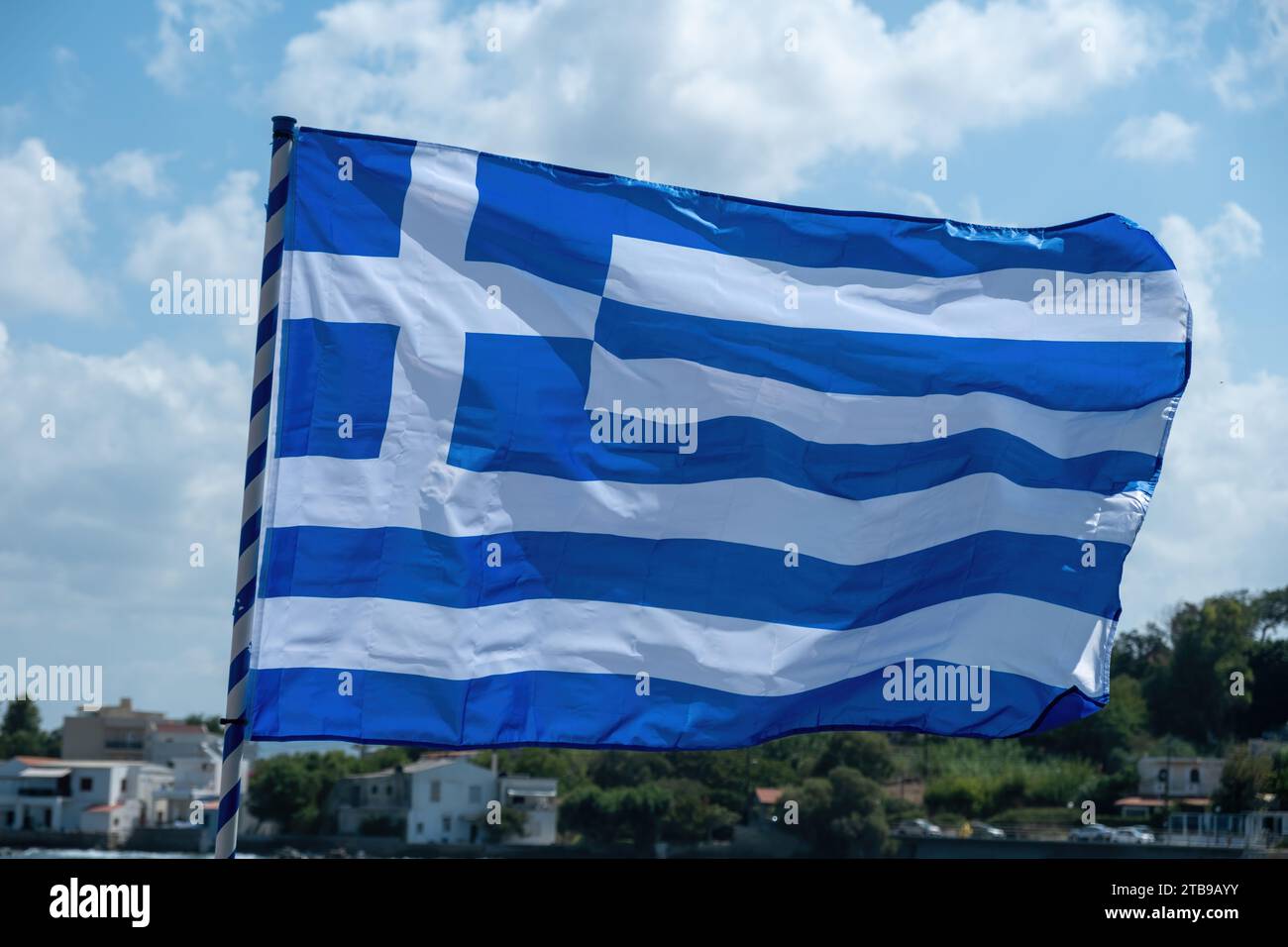 Greek flag blue and white color on flagpole waving in the wind in front of blur island, nature, building background. Greece sign symbol. Stock Photo