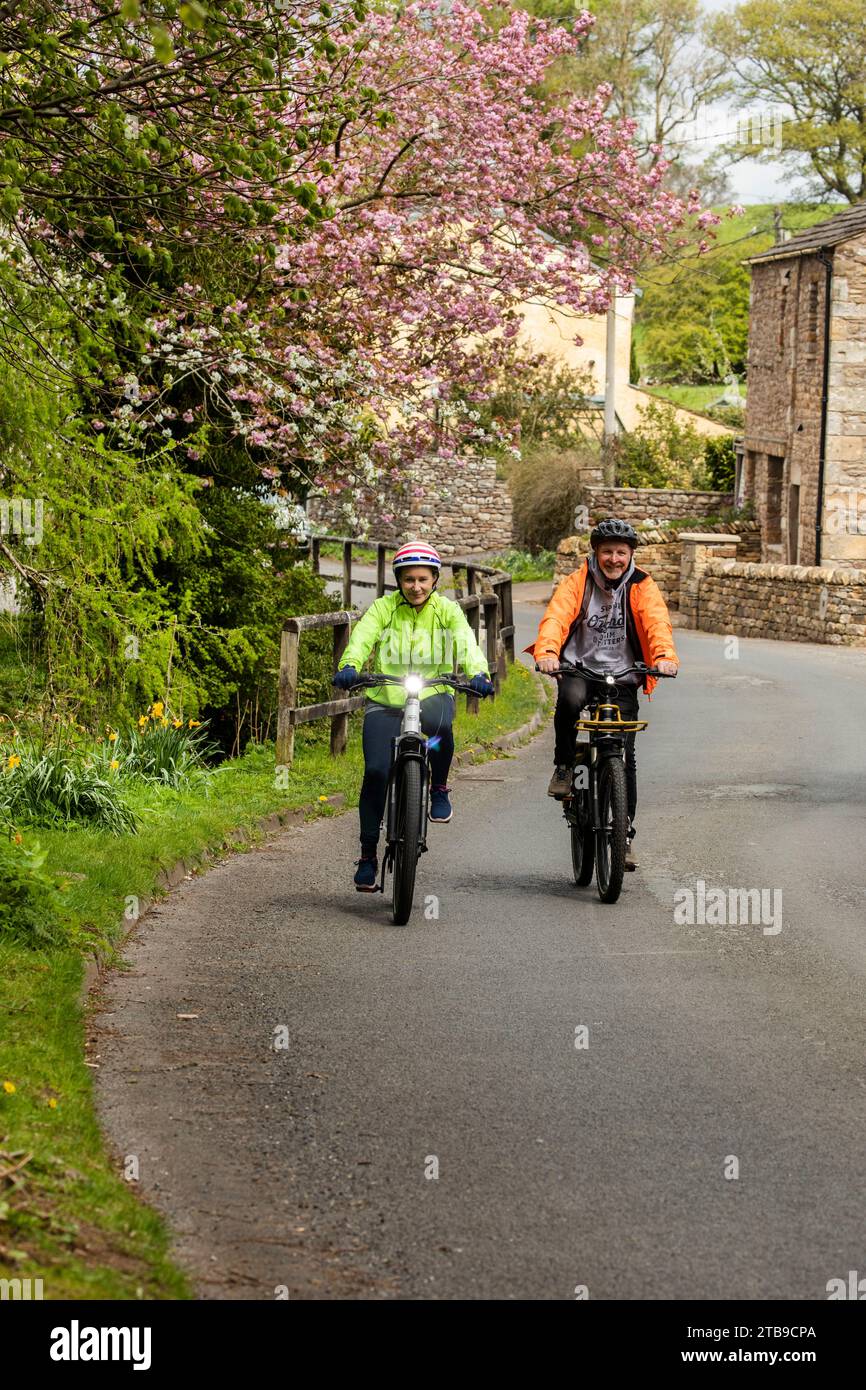 Man and woman cyclists riding electric e bikes in a pretty village in springtime, Hartley. Kirkby Stephen, North Pennines, Cumbria Stock Photo