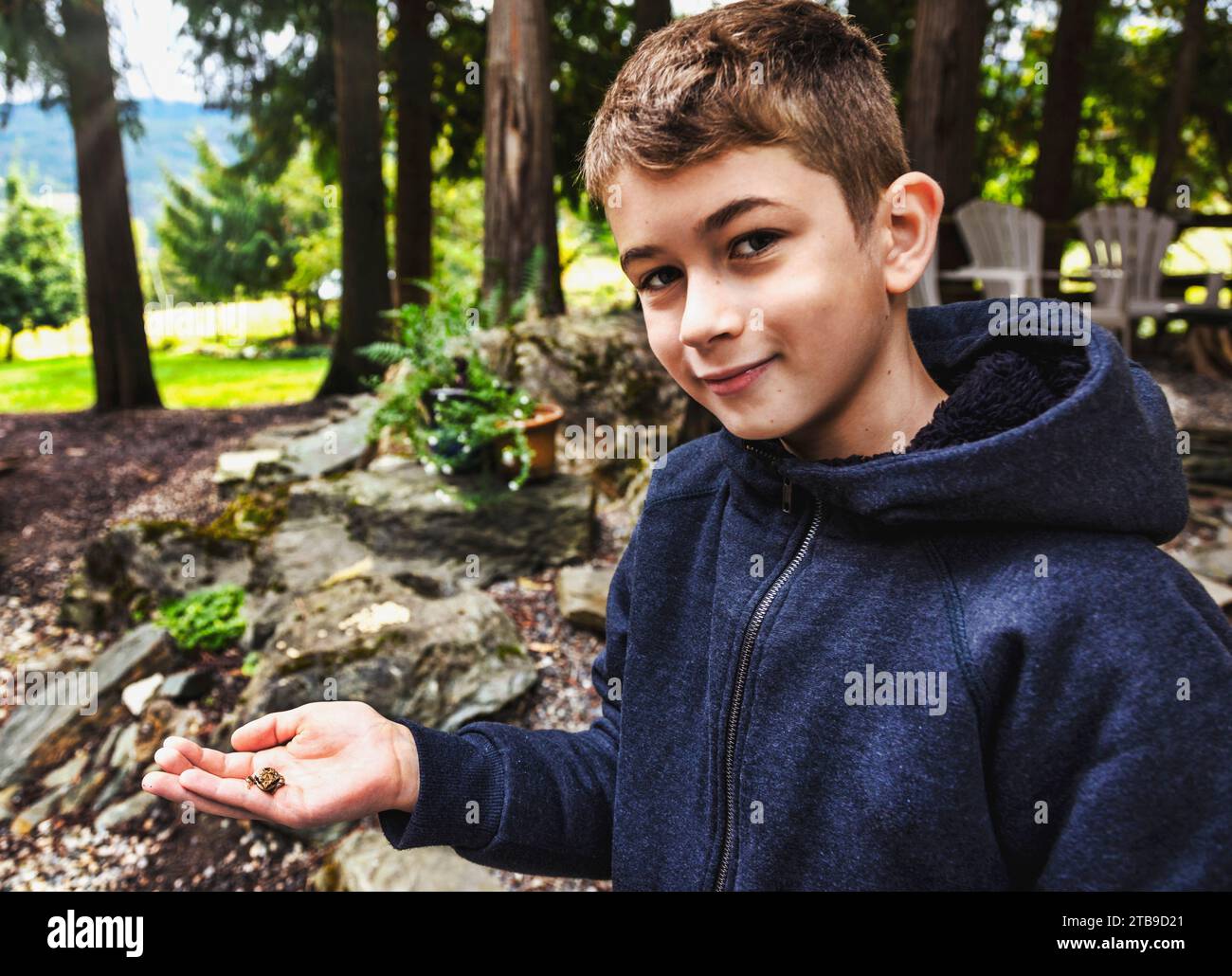 Close-up portrait of a young boy smiling at the camera and holding a small frog in his hand while on vacation; Sicamous, British Columbia, Canada Stock Photo