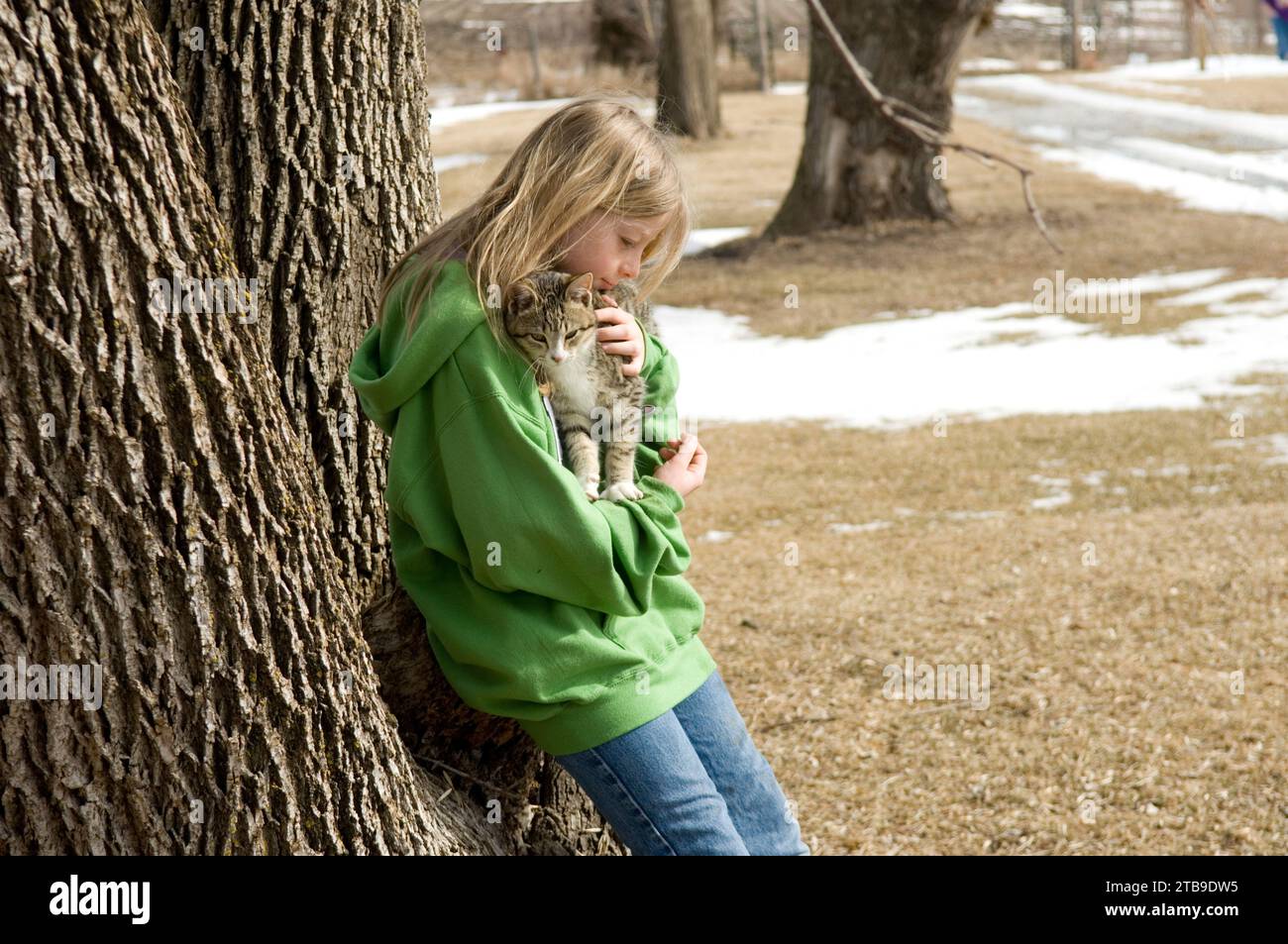 Girl stands outside and leans against a large tree as she shows affection to her pet cat; Otoe, Nebraska, United States of America Stock Photo