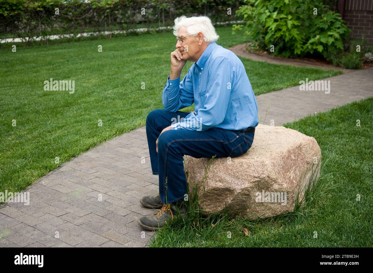 Elderly man sits in a contemplative pose on a rock in his front yard; Lincoln, Nebraska, United States of America Stock Photo