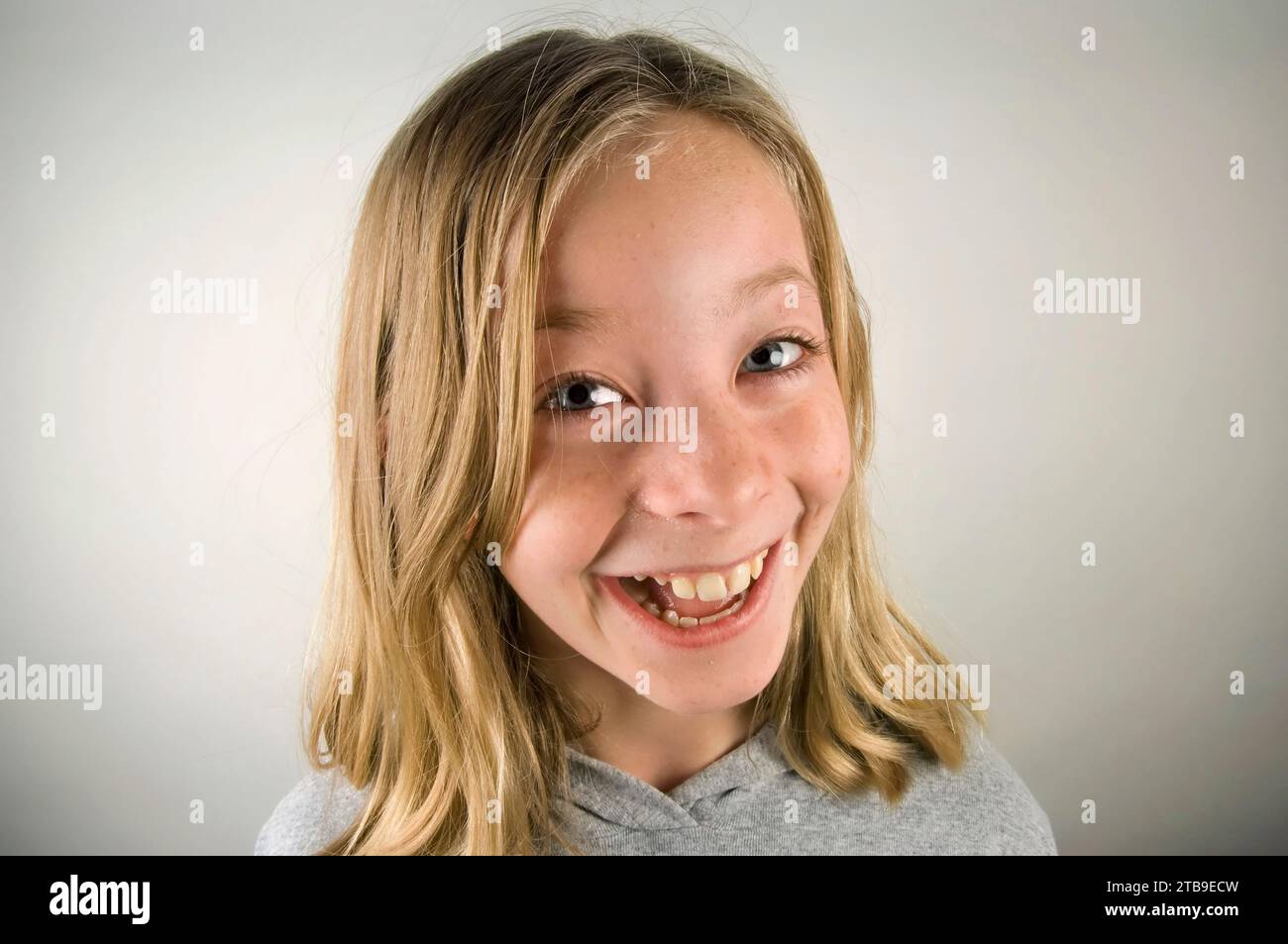 Portrait of a young girl giving a big smile to the camera, with blond hair and blue eyes against a grey background; Studio Stock Photo